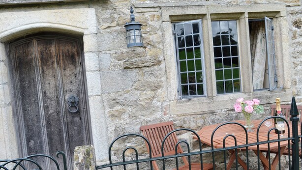 A stone building with a wooden door, a window with a grid pattern, and a table with chairs outside.