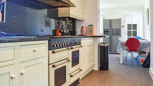 Cozinha com fogão, forno e armários. Há uma lixeira e uma mesa com cadeiras ao fundo.