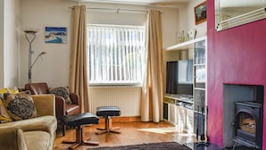 Sala de estar com sofá, poltrona, banquinho, televisão, lareira e janela com cortinas.