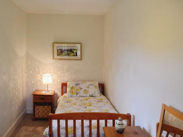 Un dormitorio con una cama, una mesita de noche con lámpara, una pequeña silla de madera y un cuadro en la pared.