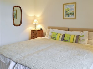 Een net opgemaakt bed met een wit geborduurd dekbed, een bedlampje, een klein houten nachtkastje en een lijst met een schilderij aan de muur.