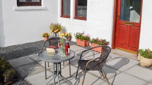A small outdoor seating area with a round table and two wicker chairs, surrounded by potted plants and a red door. - Beuchan Cottage Keir Mill (Keir Mill, Thornhill)