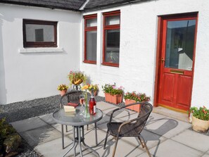 Een klein terras met een ronde tafel en twee rieten stoelen, omringd door potplanten en een rode deur.