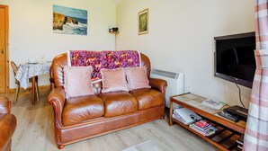 Una sala de estar con un sofá de cuero marrón, una mesa de comedor con sillas, un televisor y una mesita con libros y un teclado.