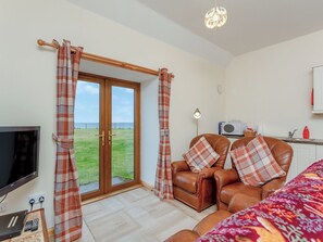 A room with a large sliding glass door to a grassy area, a leather sofa with plaid cushions, a television, and a small kitchenette.
