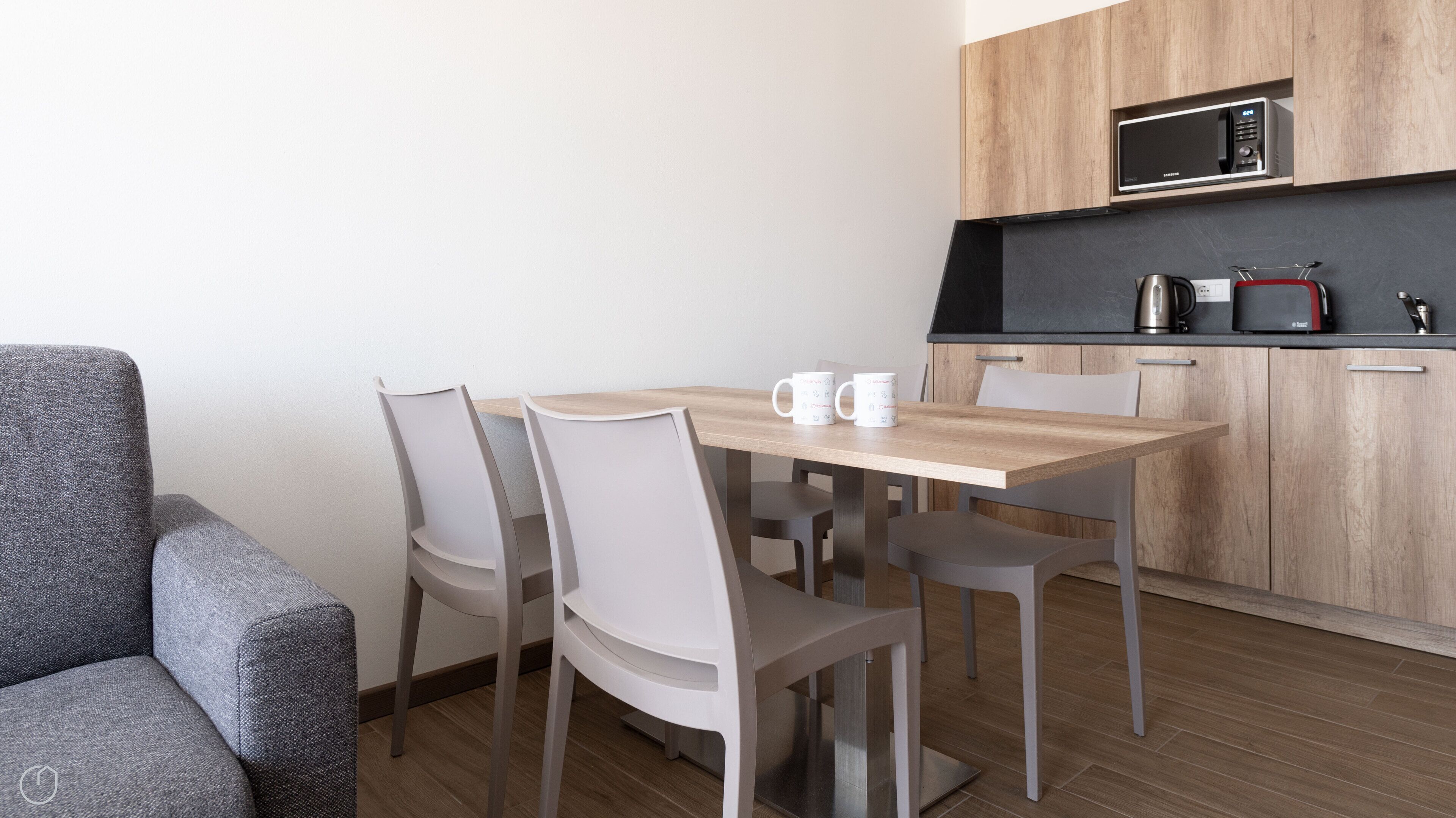 Een moderne keuken met een magnetron, koffiezetapparaat en broodrooster. Er is een eettafel met vier stoelen en een grijze bank.