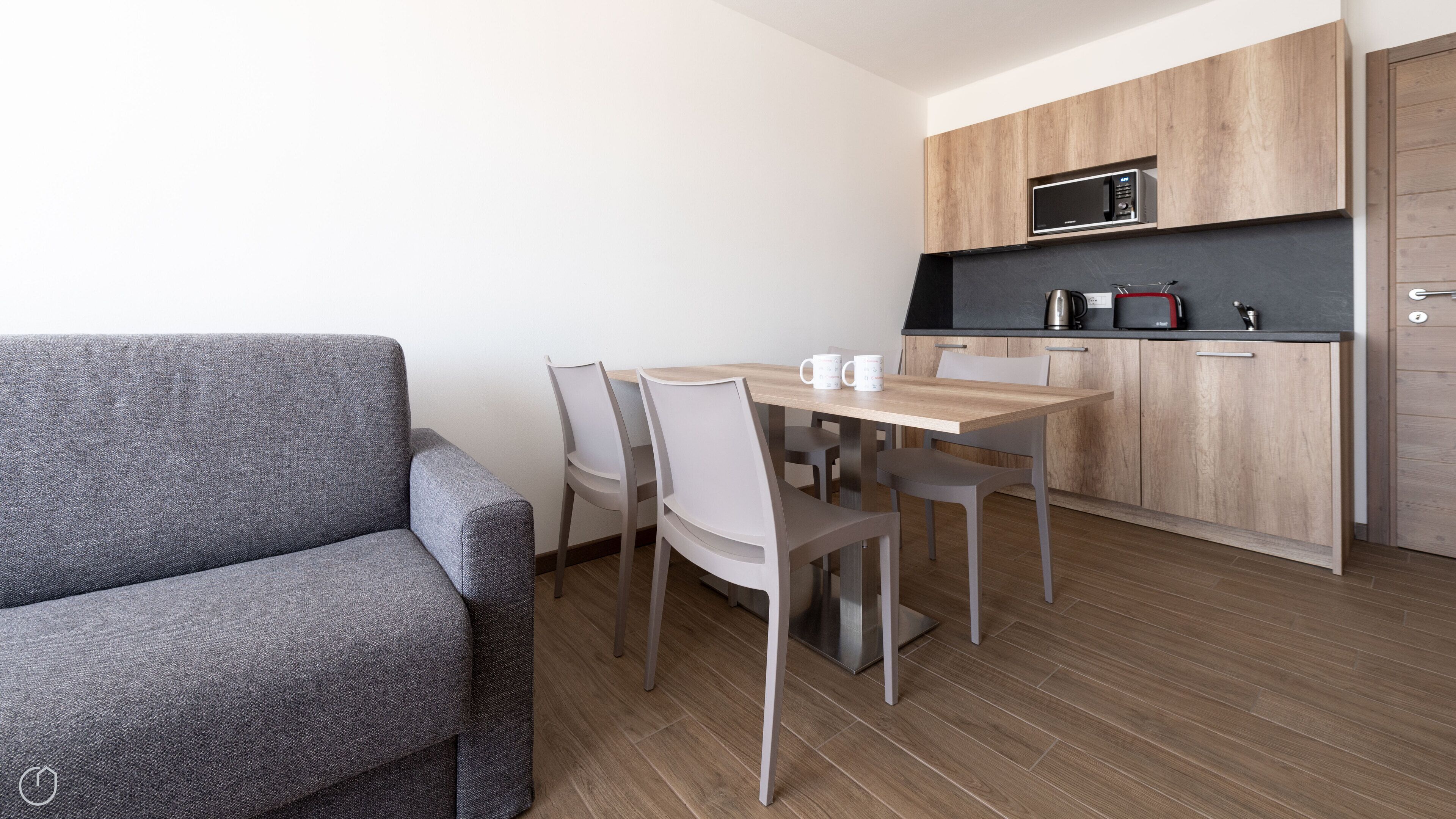 Un espace de vie compact comprenant un canapé gris, une table à manger en bois et une kitchenette équipée d’un four à micro-ondes.