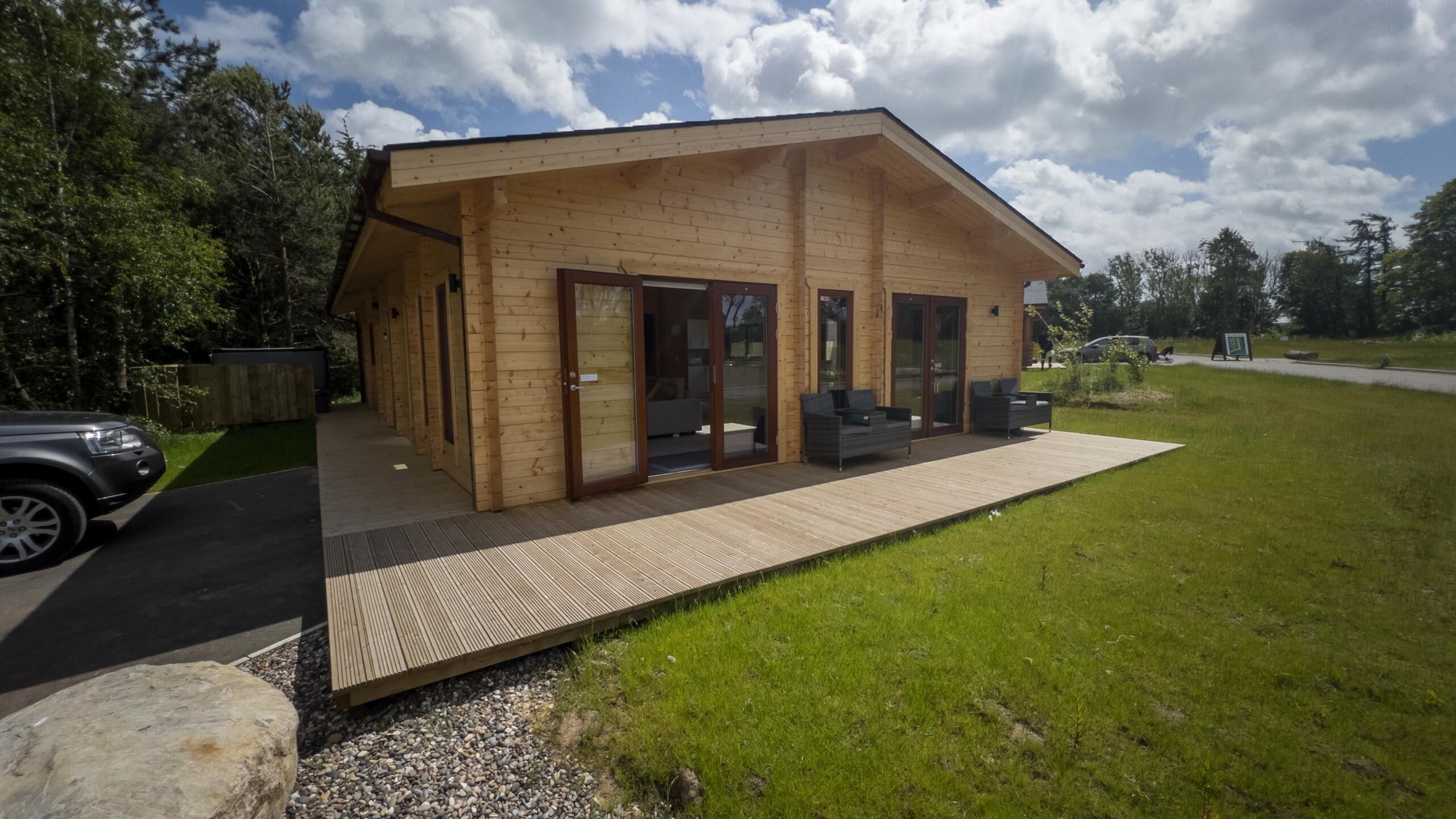 A wooden cabin with a covered patio, a car parked in front, and a grassy area.