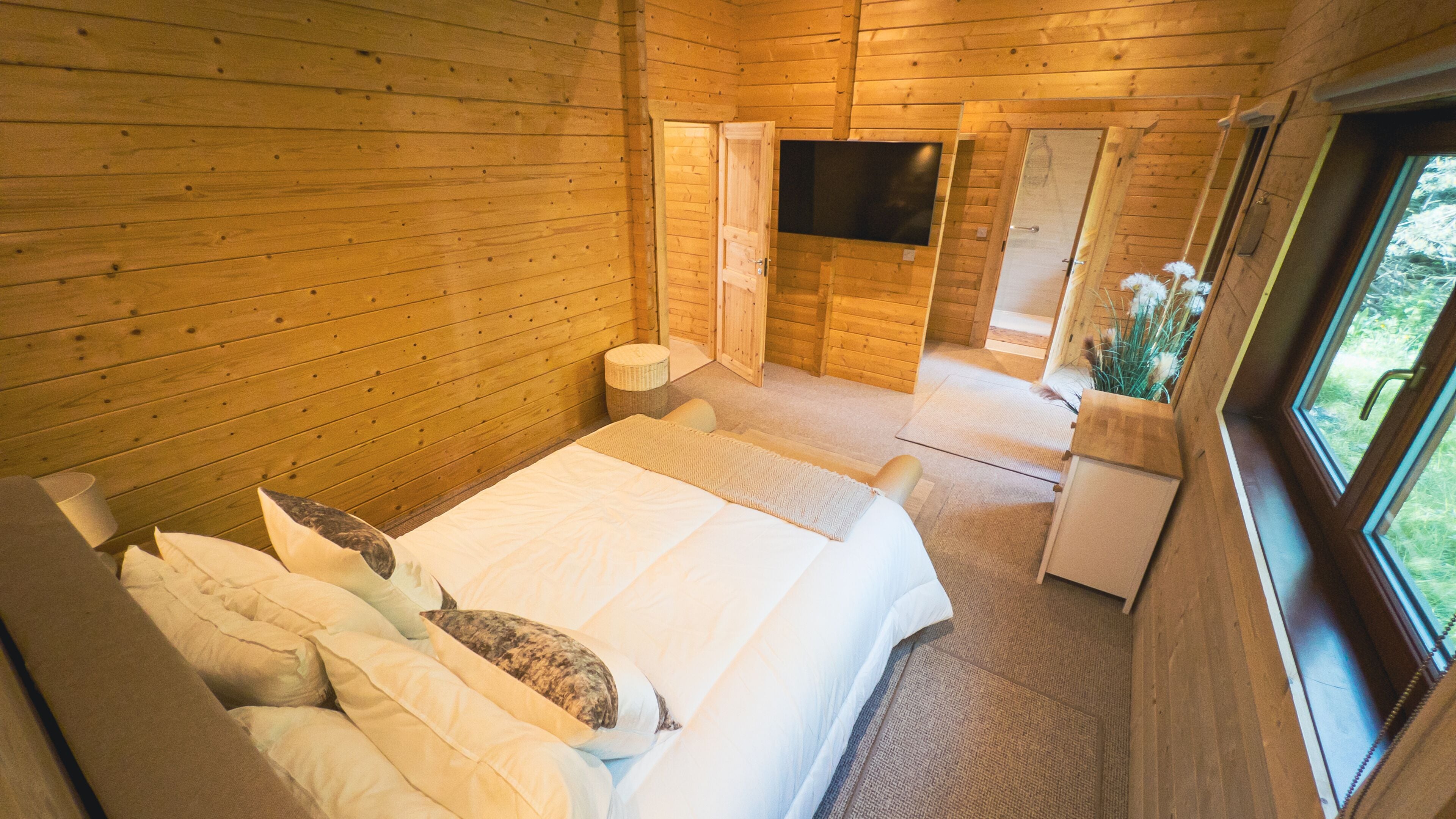 Quarto de cabana de madeira com uma cama, travesseiros, uma pequena mesa de cabeceira, televisão e porta do banheiro.