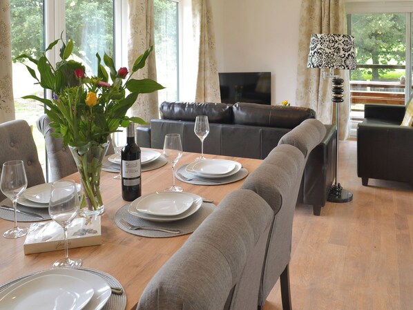 A dining area with a wooden table set for four, surrounded by grey upholstered chairs. A vase with flowers is on the table, and a bottle of wine is visible. A television is mounted on the wall in the background.