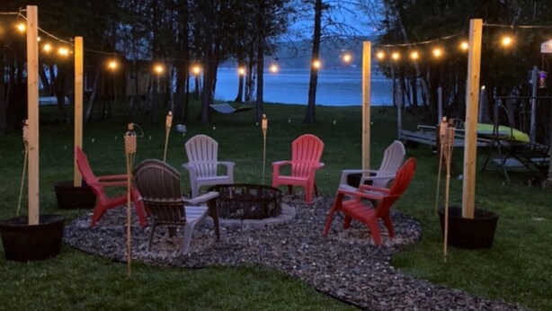 An outdoor seating area with Adirondack chairs and a fire pit, surrounded by string lights and a view of the lake. - Newly Renovated, perfectly located and fully equiped. (Portage Lake)