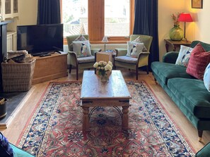 Un salon comprenant une télévision, deux fauteuils, un canapé, une table basse ornée de fleurs, un tapis à motifs et une fenêtre avec des rideaux.