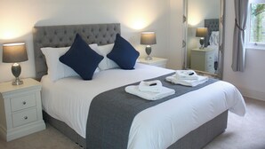 Una cama bien hecha con dos almohadas, una mesita de noche con una lámpara, un espejo y una ventana con cortinas.