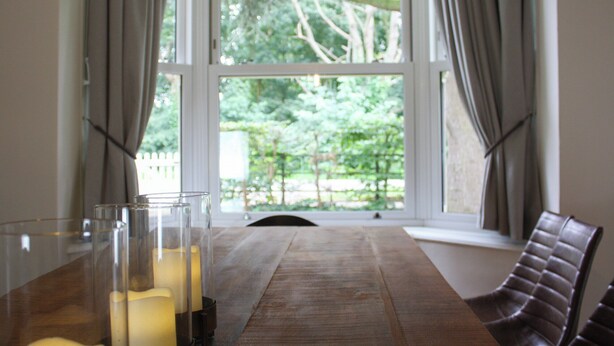 Un comedor con una mesa de madera, candelabros de vidrio transparente y vistas a árboles a través de amplias ventanas.