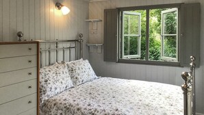 A bedroom with a bed, a dresser, a window with a view of greenery, and a wall-mounted light fixture. - Fig Tree Shepherds Hut (Bridport)