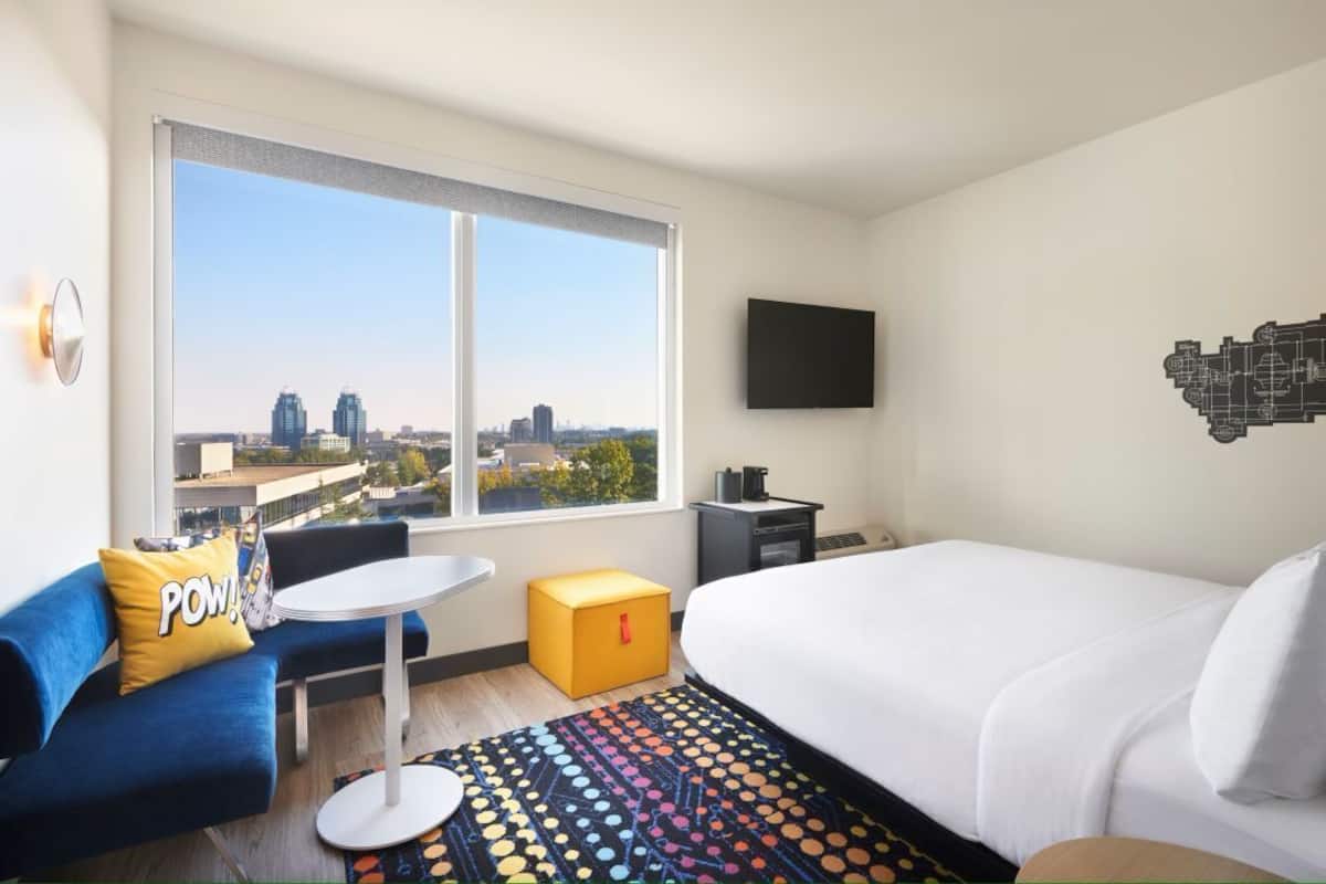 A hotel room with a large window, a bed, a desk, a chair, and a colorful rug.
