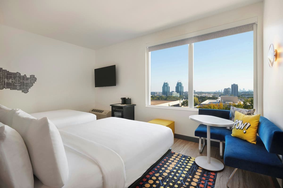 A modern hotel room with a large window offering a city view, a bed with white linens, a blue sofa with a yellow pillow, and a small round table.