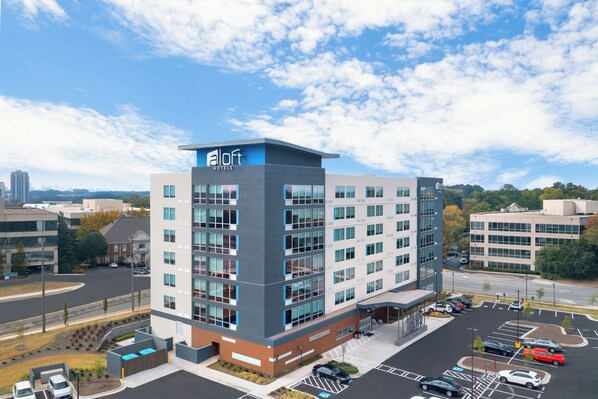 A modern hotel building with a flat roof, multiple floors, and a parking lot in front.