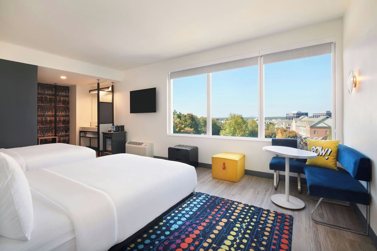 A hotel room with a large bed, a sofa, a small table, a TV, a bookshelf, and a window with a city view.