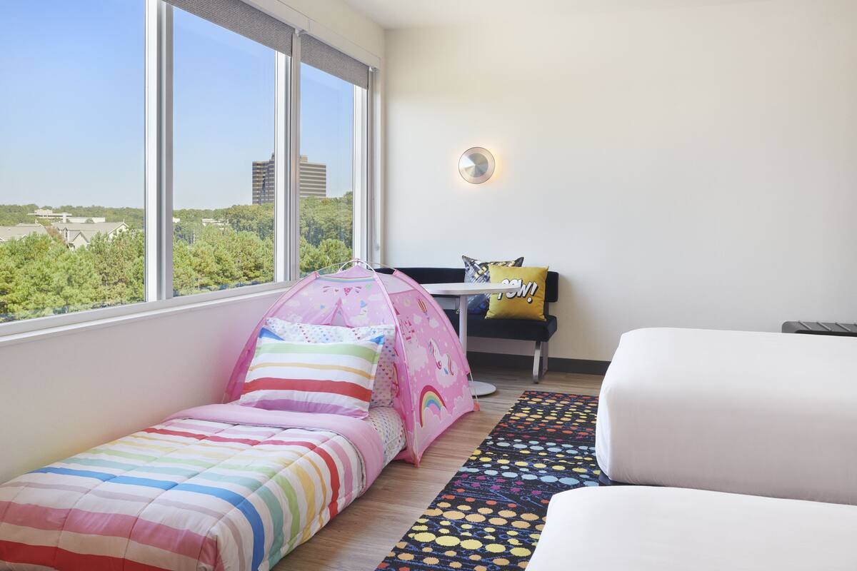 A bedroom with a bed, a desk, and a window with a view of buildings and trees.