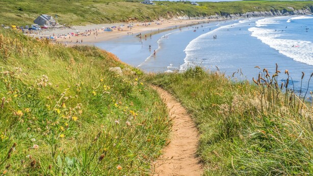Ein Küstenpfad, der zu einem Strand mit grünen Flächen und einigen Gebäuden im Hintergrund führt.