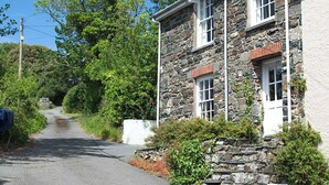 Ein Steinhaus mit weißer Tür und Fenstern, umgeben von Grünpflanzen und einer schmalen Straße.