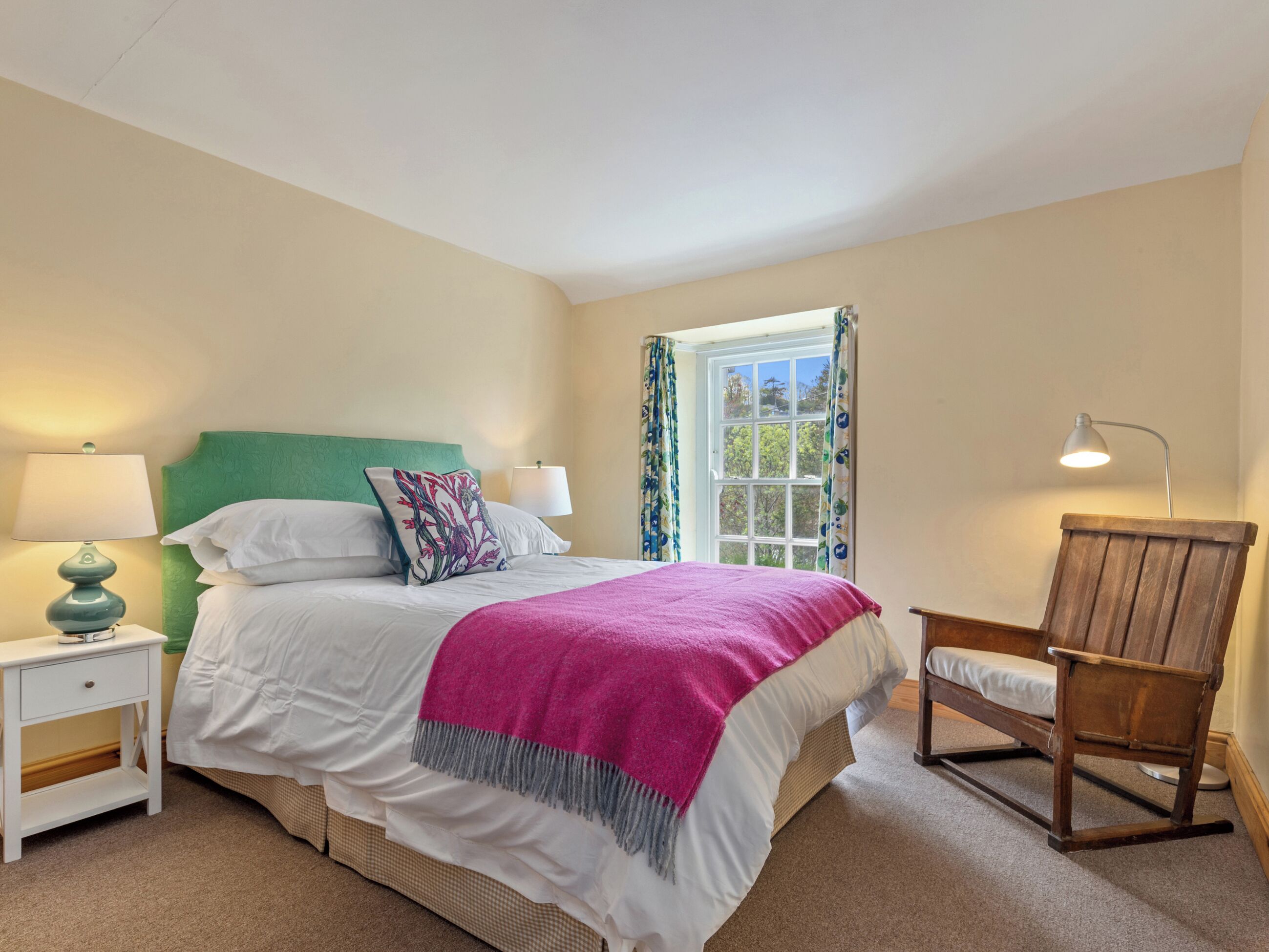 A bedroom with a bed, a nightstand, a chair, a window with curtains, and a lamp.