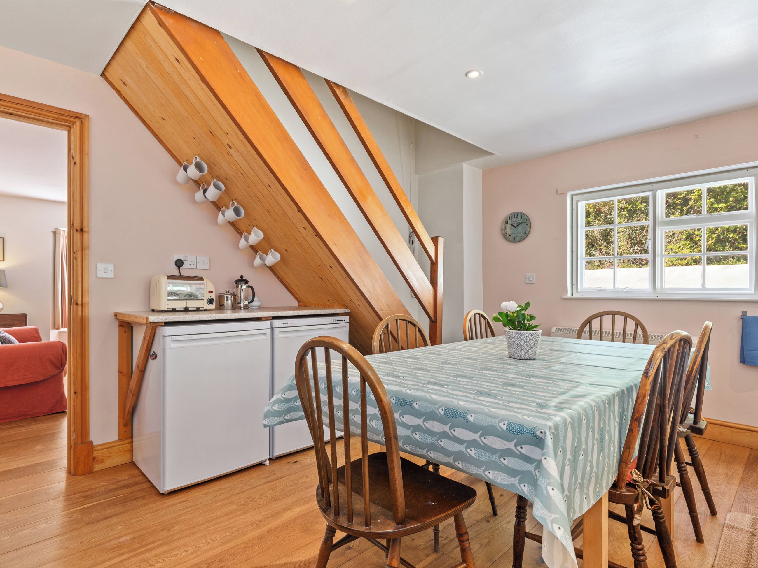 A dining area with a table set for four, wooden chairs, a kitchen area with a refrigerator, and a window with a view of greenery.