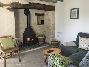 Una sala de estar acogedora con chimenea de piedra, una silla de madera con cojín verde, un sofá gris con cojines estampados y una cesta tejida sobre un taburete de madera.
