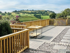 Una terraza de madera con una mesa y sillas, rodeada por un camino de piedra y vistas a colinas ondulantes.
