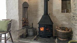 Ein rustikaler Kamin aus Stein mit einem schwarzen Ofen, ein Holzstuhl und ein Holzkorb mit Brennholz.