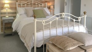 Ein Schlafzimmer mit weißem Metallbettgestell, einem Holzkopfteil, einem Nachttisch mit Lampe und einer hölzernen Kommode.