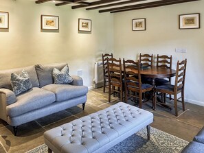 Ein Wohnzimmer mit grauem Sofa, einem gesteppten Hocker, einem Esstisch mit Holzstühlen und gerahmten Bildern an der Wand.