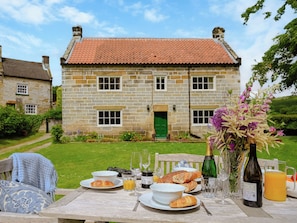 Una casa de piedra con tejado de baldosas, una mesa de comedor dispuesta con comida y bebidas, y un jardín al fondo.