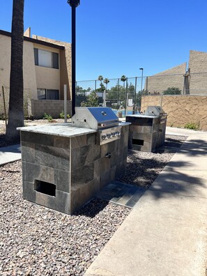 Dos parrillas para barbacoa al aire libre con bases de piedra, frente a un cielo azul despejado, palmeras y un edificio con fachada texturizada.