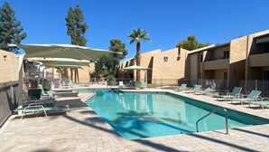 Ein Poolbereich mit Liegestühlen und Sonnenschirmen, umgeben von Gebäuden mit heller Fassade.