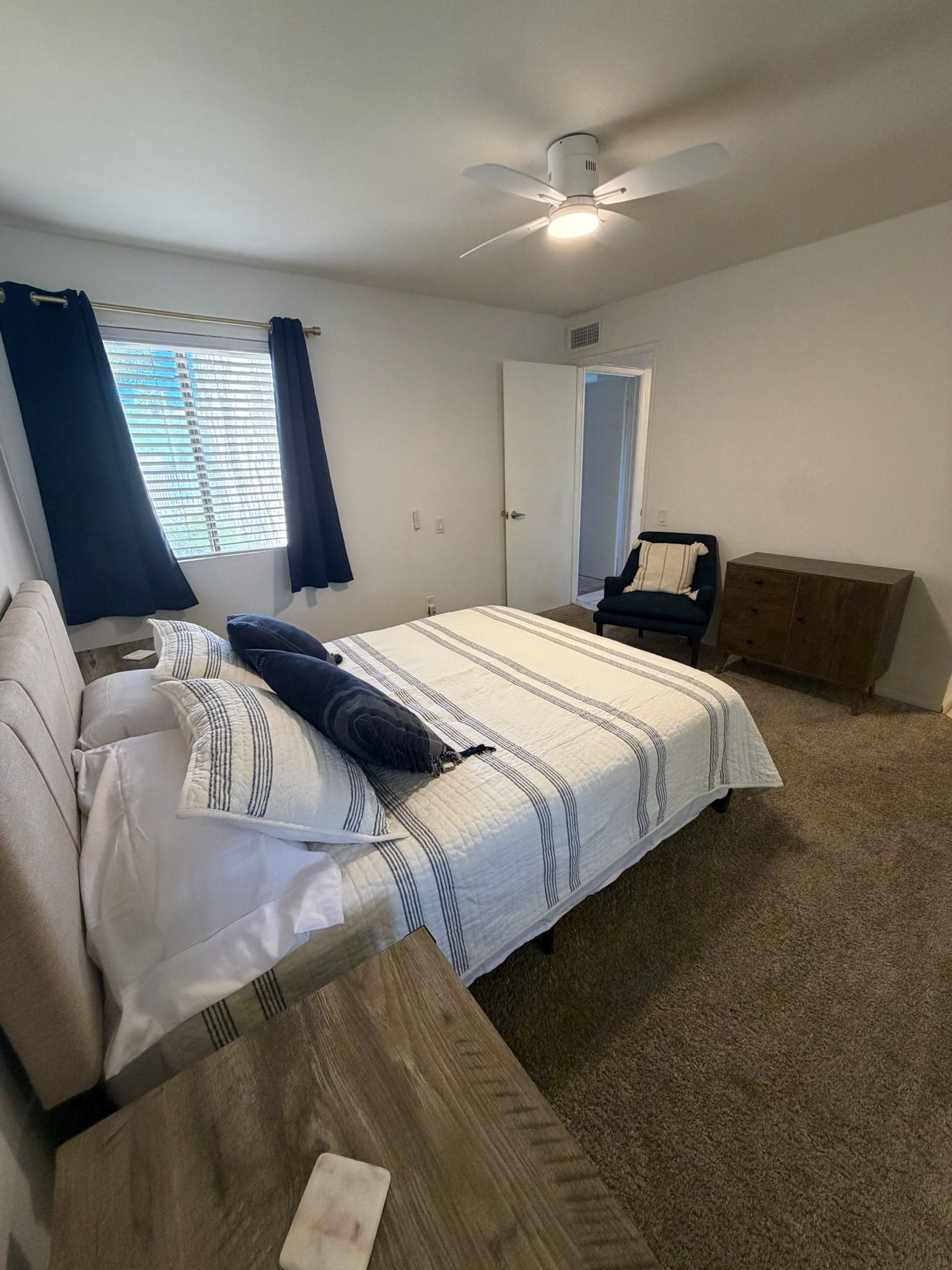 Une chambre à coucher comprenant un lit, une chaise, un bureau, un ventilateur de plafond et une fenêtre avec des rideaux.