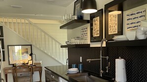 A modern kitchen with white cabinets, a black countertop, and a herringbone backsplash. There is a dining area with a wicker chair and a framed picture on the wall. - "Bee Keeper's Cottage" on private estate, new construction, close to everything! (Sarasota)