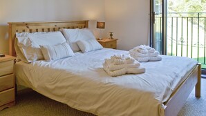 A neatly made bed with white linens and pillows, a bedside table with a lamp, and a view of greenery through a balcony door.