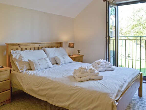 A neatly made bed with white linens and pillows, a bedside table with a lamp, and a view of greenery through a balcony door.