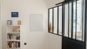 A room with a bookshelf, a framed picture, and a window with a view of a building. - Le Saint Firmin - Two Bedroom Apartment, Sleeps 4 (Montpellier)