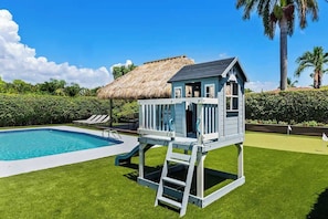Zwembad met een blauw dek, een speelhuisje en een constructie met rieten dak.