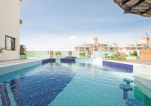 A swimming pool with clear blue water, surrounded by a tiled deck and a low wall. - Apartamento Classe A (Florianópolis)