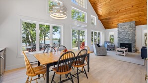 A spacious dining area with a wooden table and chairs, large windows, a stone fireplace, and a view of the outdoors. - Water Front Cottage in Stanley Bridge- Just Minutes To Cavendish Beach (Breadalbane)