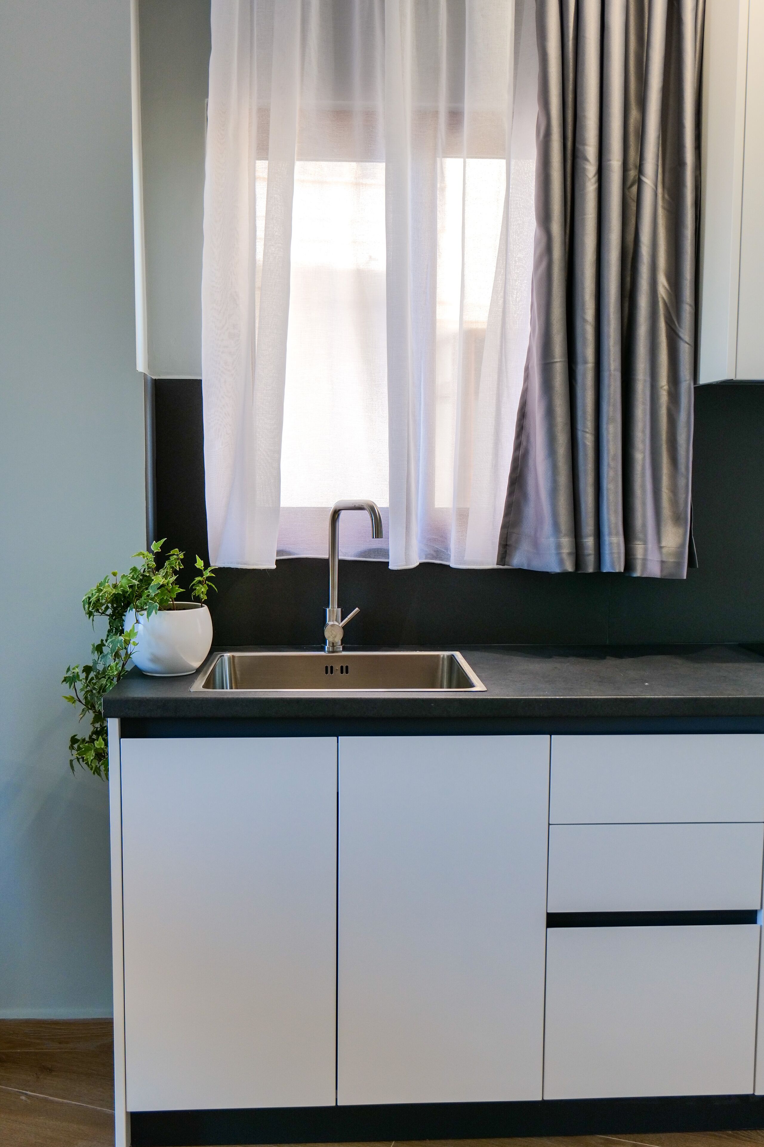 A modern kitchen with a stainless steel sink, white cabinets, and a window with sheer curtains.