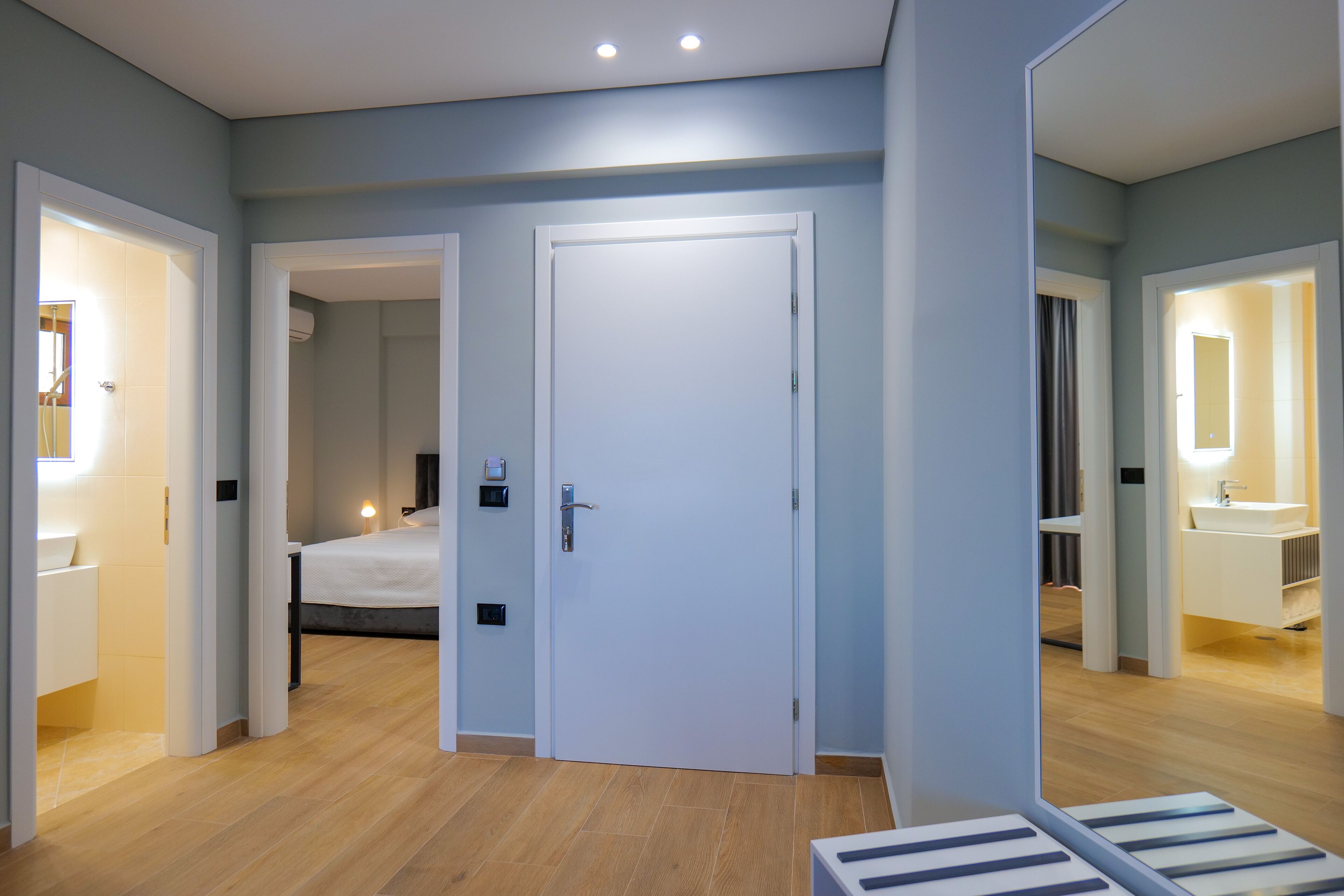 Une chambre d’hôtel moderne avec un plancher en bois, un lit, une salle de bain équipée d’un lavabo et d’un miroir, et une porte donnant sur une autre pièce.