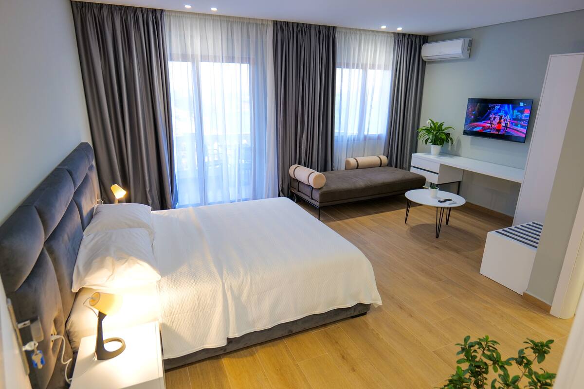 A modern hotel room with a large bed, a sofa, a small table, a TV, and a potted plant.