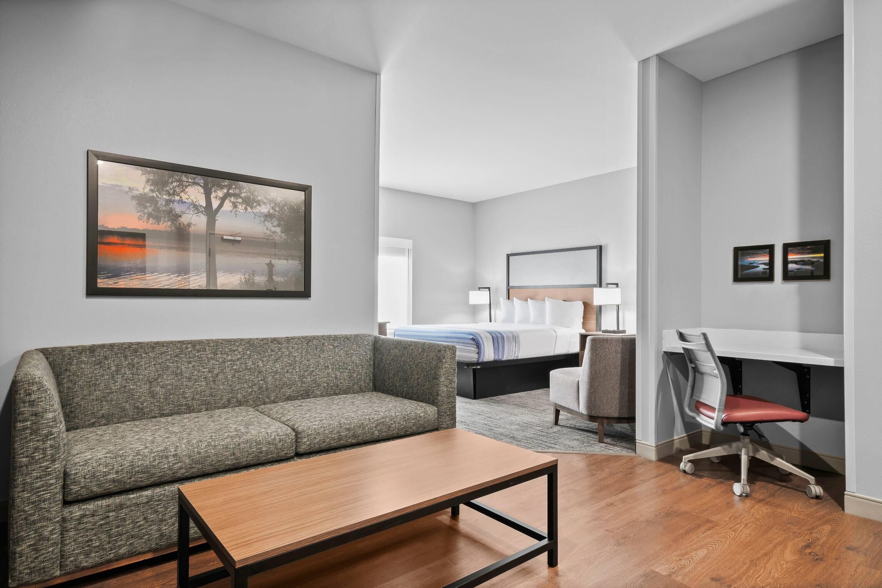 A modern hotel room with a grey sofa, a wooden coffee table, a desk with a chair, a bed with white linens, and a painting of a tree by a lake.