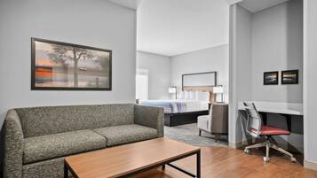 A modern hotel room with a grey sofa, a wooden coffee table, a desk with a chair, a bed with white linens, and a painting of a tree by a lake.
