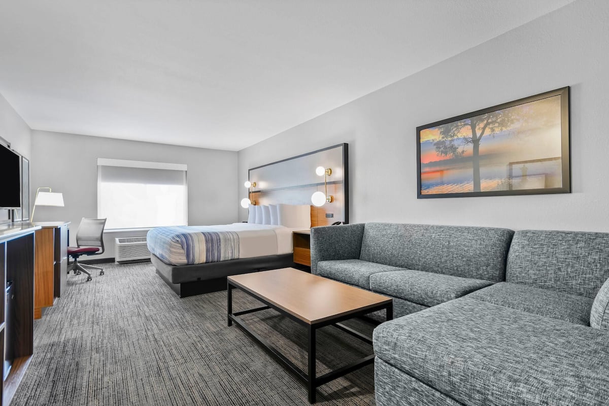 Ein modernes Hotelzimmer mit einem Bett, einem grauen Sofa, einem Holztisch, einem Schreibtisch mit Computer und einem großen Fenster mit Vorhängen.